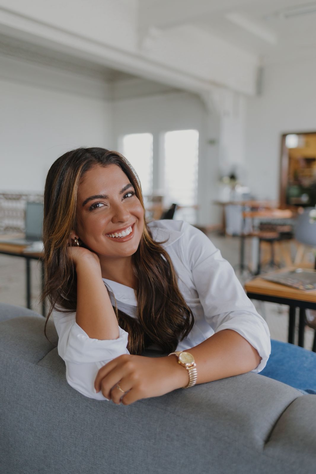 Amanda Prado, psicóloga, sorrindo apoiada em um sofá no consultório
